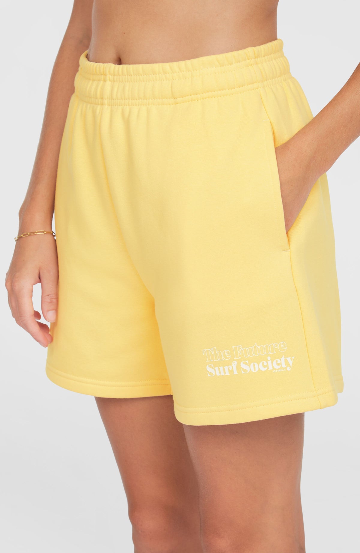 Nahaufnahme der gelben Future Surf Society Shorts für Damen am Model, Fokus auf Seitentasche und Logo-Print am Saum