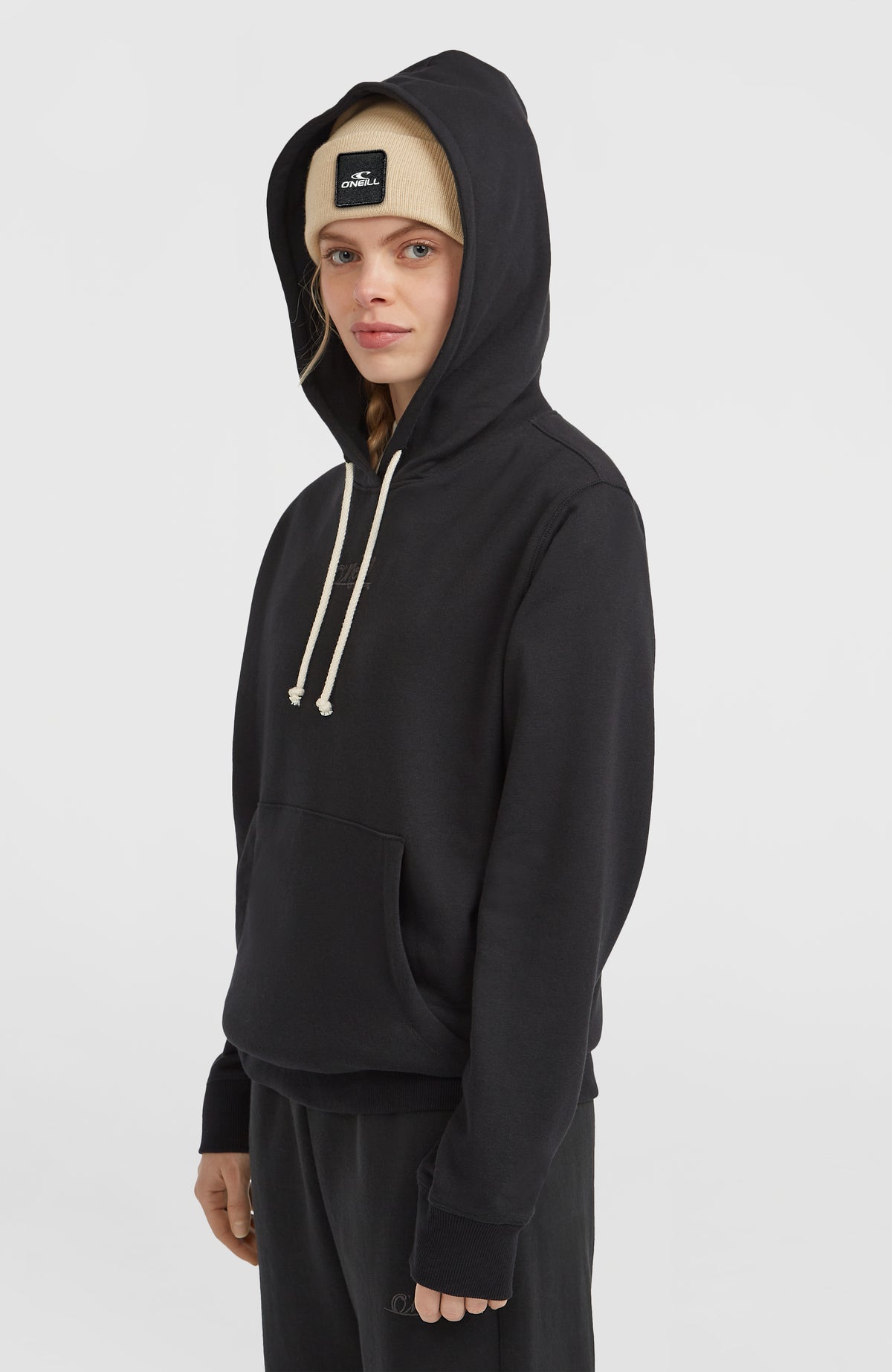 Essentials Script Hoodie für Damen, schwarz, Halbprofil getragen am Model mit aufgesetzter Kapuze und Kängurutasche