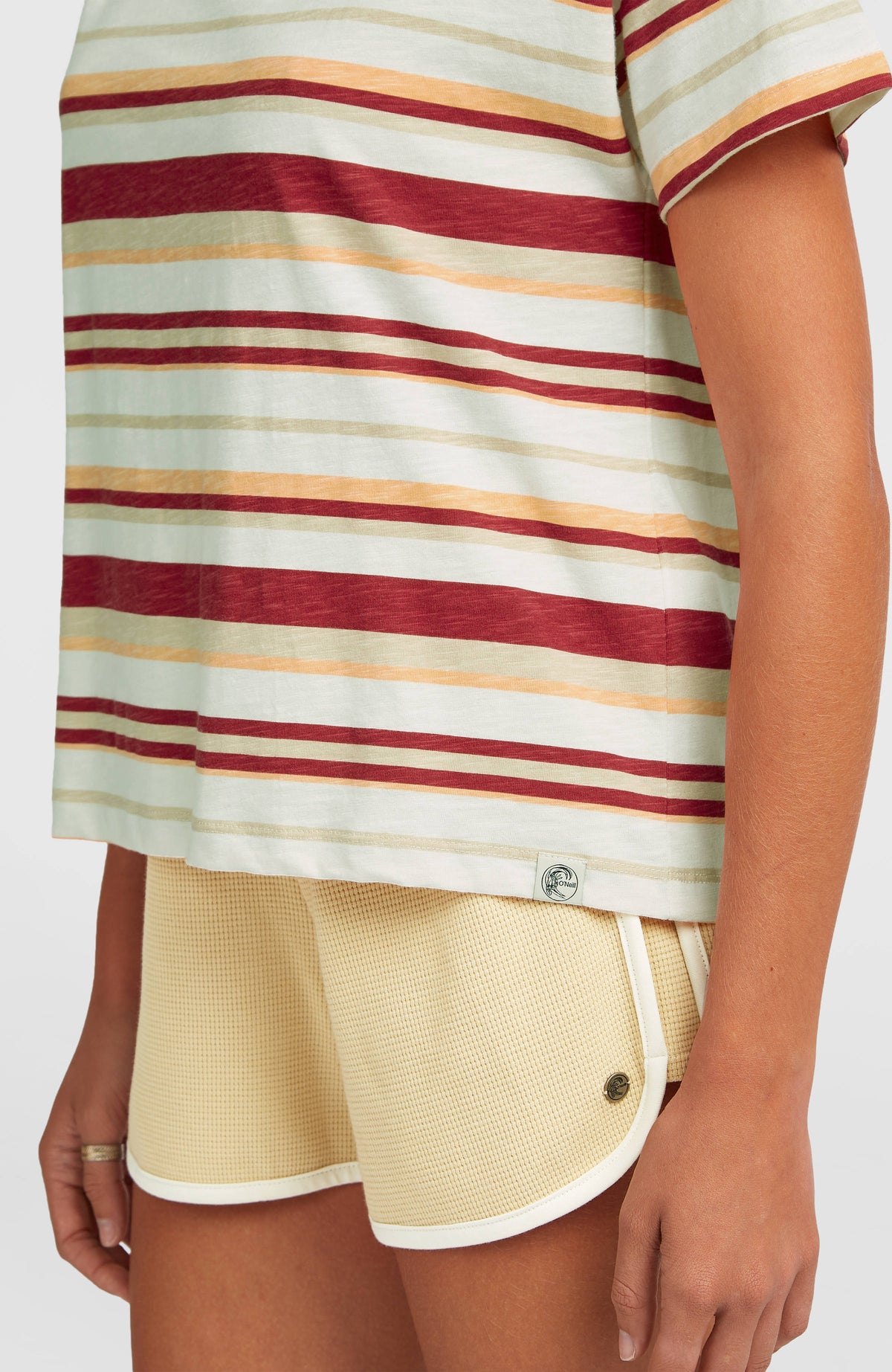 Nahaufnahme des beigen O'Riginals Boxy Stripe T-Shirt mit Streifenmuster in Rot und Orange, Logo-Badge am Saum