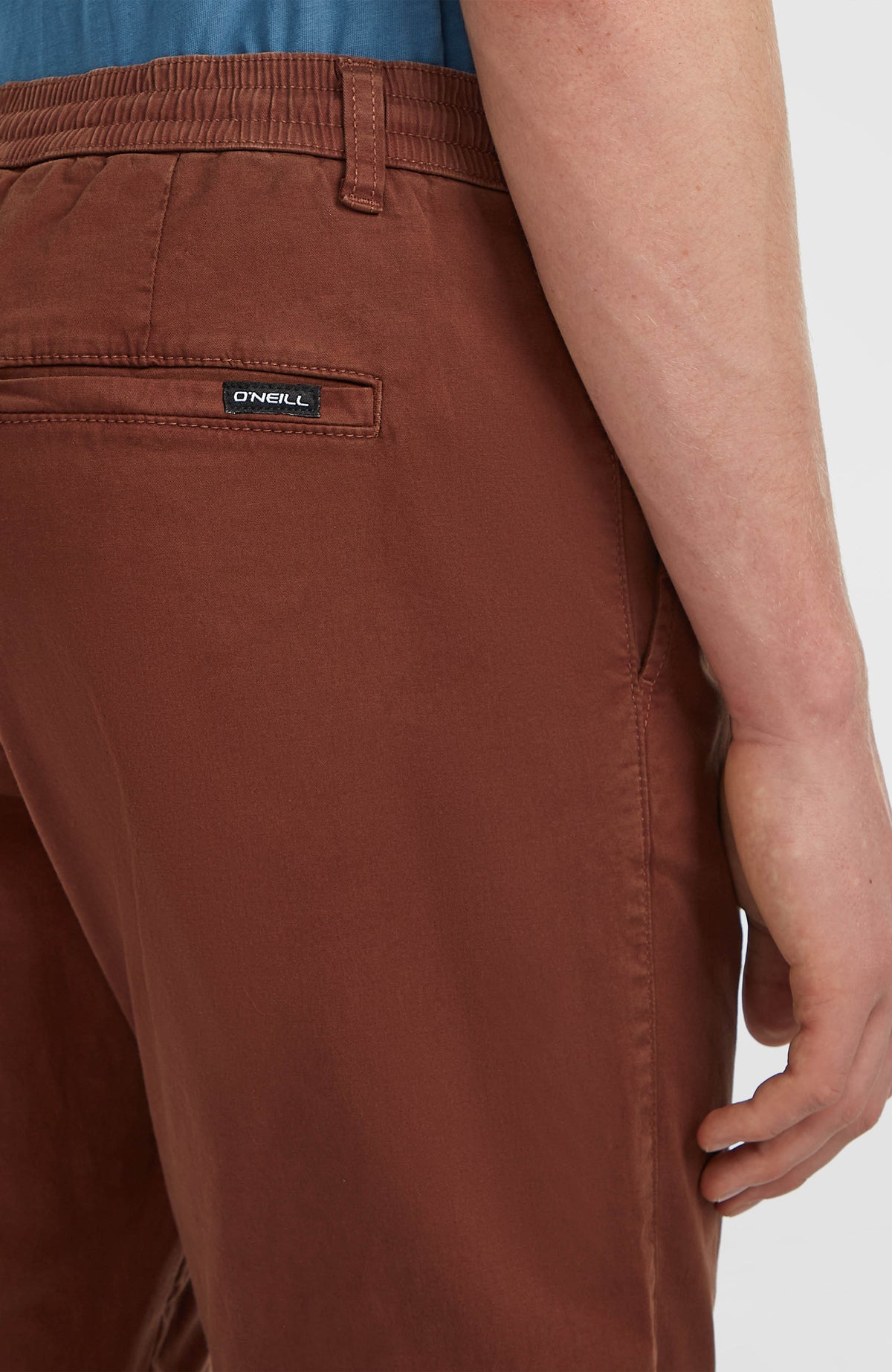 Nahaufnahme der braunen Essentials Chino-Hose am Model, Gesäßtasche mit kleinem Logo-Badge am Bund