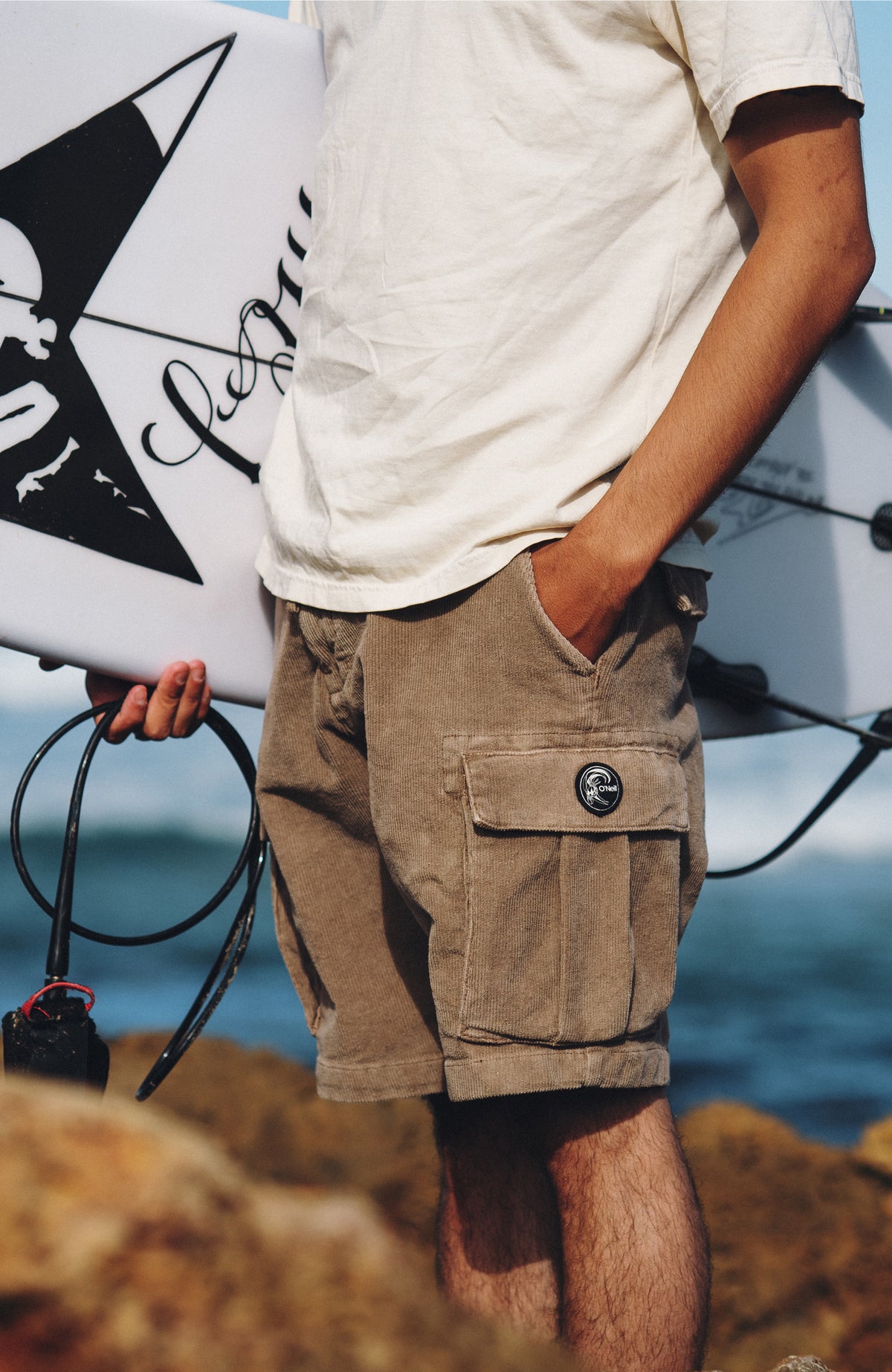 O'Riginals Cord-Cargo-Shorts für Herren, braun, getragen am Model an felsiger Küste mit Surfbrett