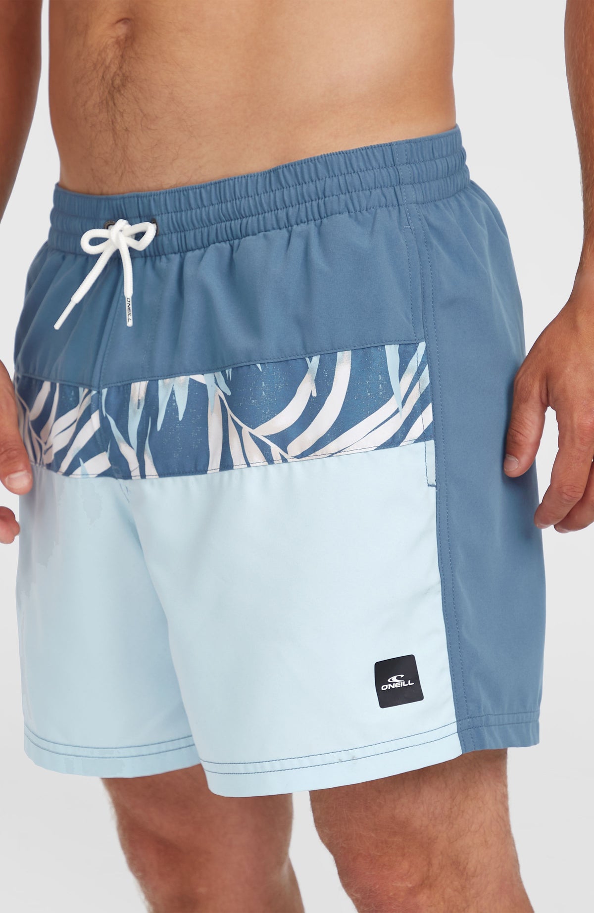 Nahaufnahme der blauen Cali Block 15'' Badehose mit Blättermuster in Weiß am Model, Kordelzug und Logo-Badge
