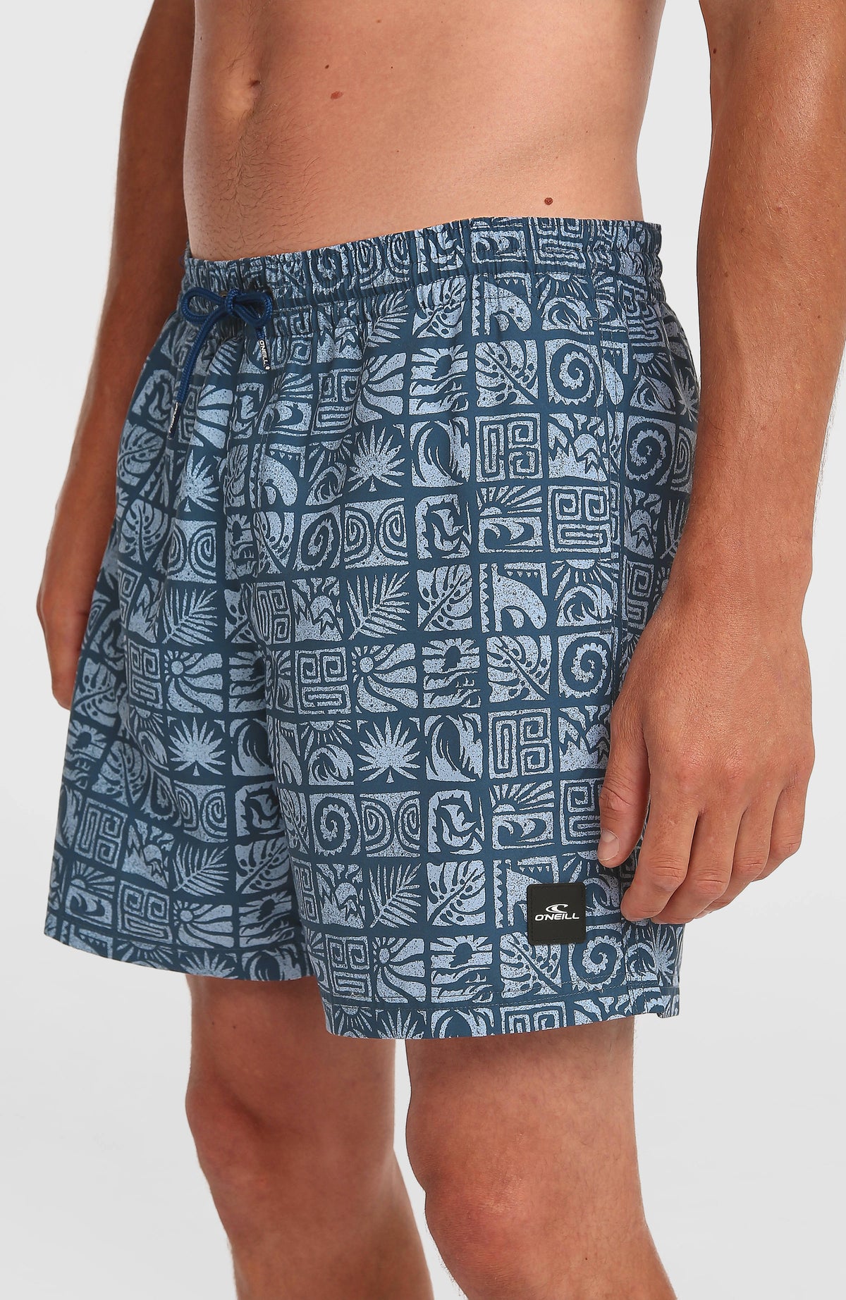 Print 16'' Badehose, blau mit Grafikmuster in Hellblau, Nahansicht am Model mit Logo-Patch am Saum