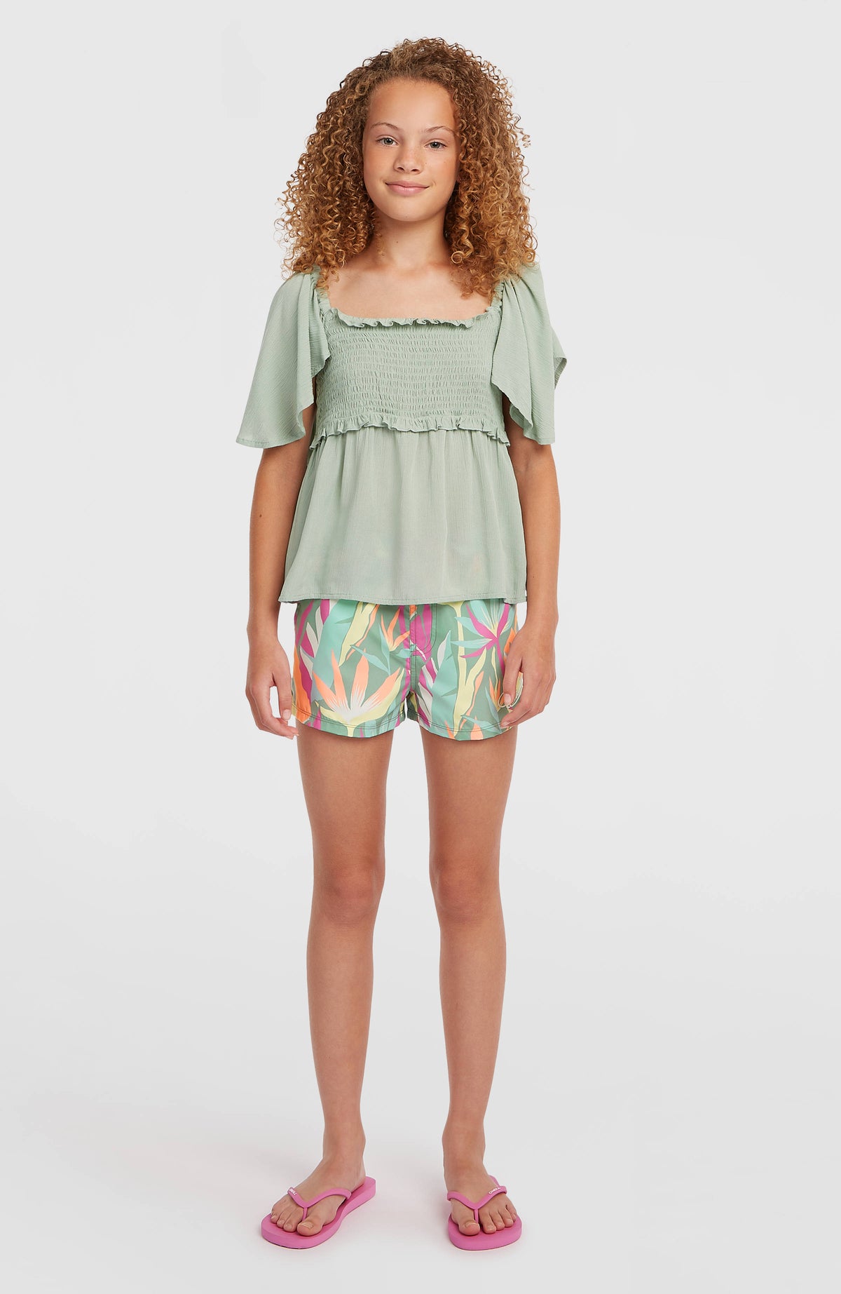 Essentials Badehose für Mädchen, grün mit Tropenmuster in Pink und Gelb, stehende Ganzkörperansicht am Model