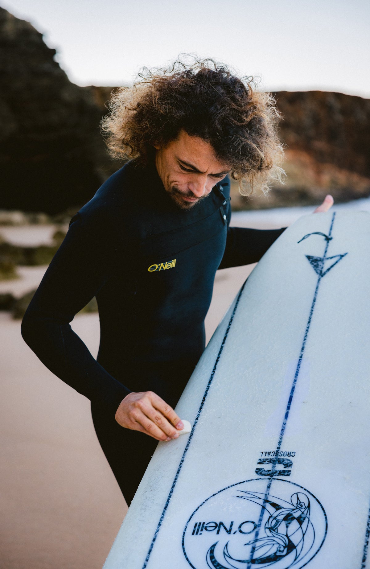 O'Riginals RG8 Neoprenanzug für Herren, schwarz, getragen am Model am Strand, Vorderansicht mit Surfbrett