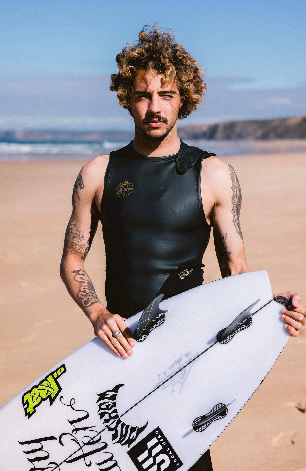 O'Riginals 2mm Long John Wetsuit Neoprenanzug für Herren, schwarz, am Strand mit Surfbrett, Vorderansicht