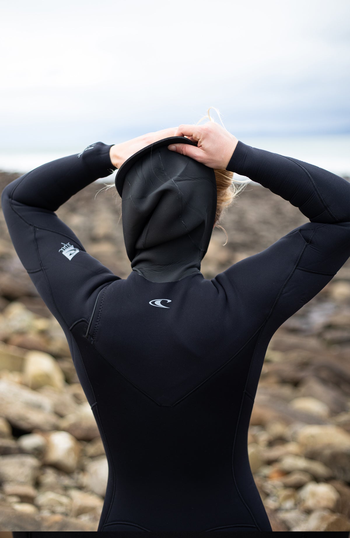 Epic 6/4 Chest Zip Wetsuit with Hood Neoprenanzug für Damen, schwarz, Rückansicht getragen am Model am steinigen Strand