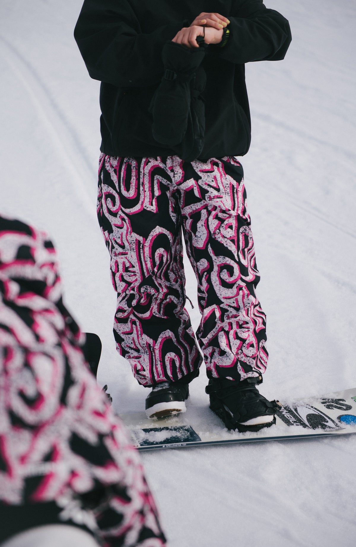FWC'Play Ski- und Snowboardhose, weiß mit Grafikmuster in Schwarz und Pink, getragen am Model im Schnee auf dem Snowboard
