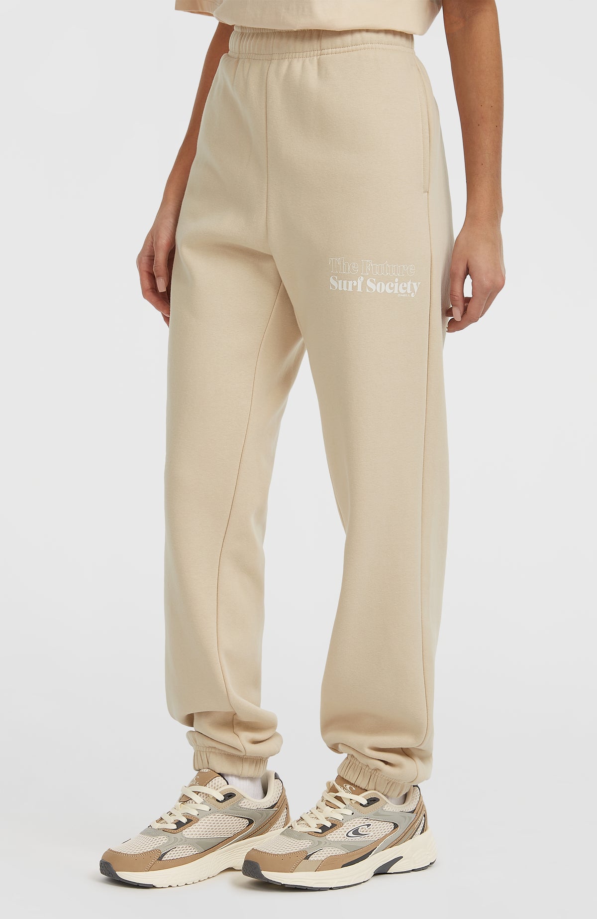 Future Surf Society Jogginghose für Damen, beige mit Logo-Print in Weiß, schräge Seitenansicht am Model
