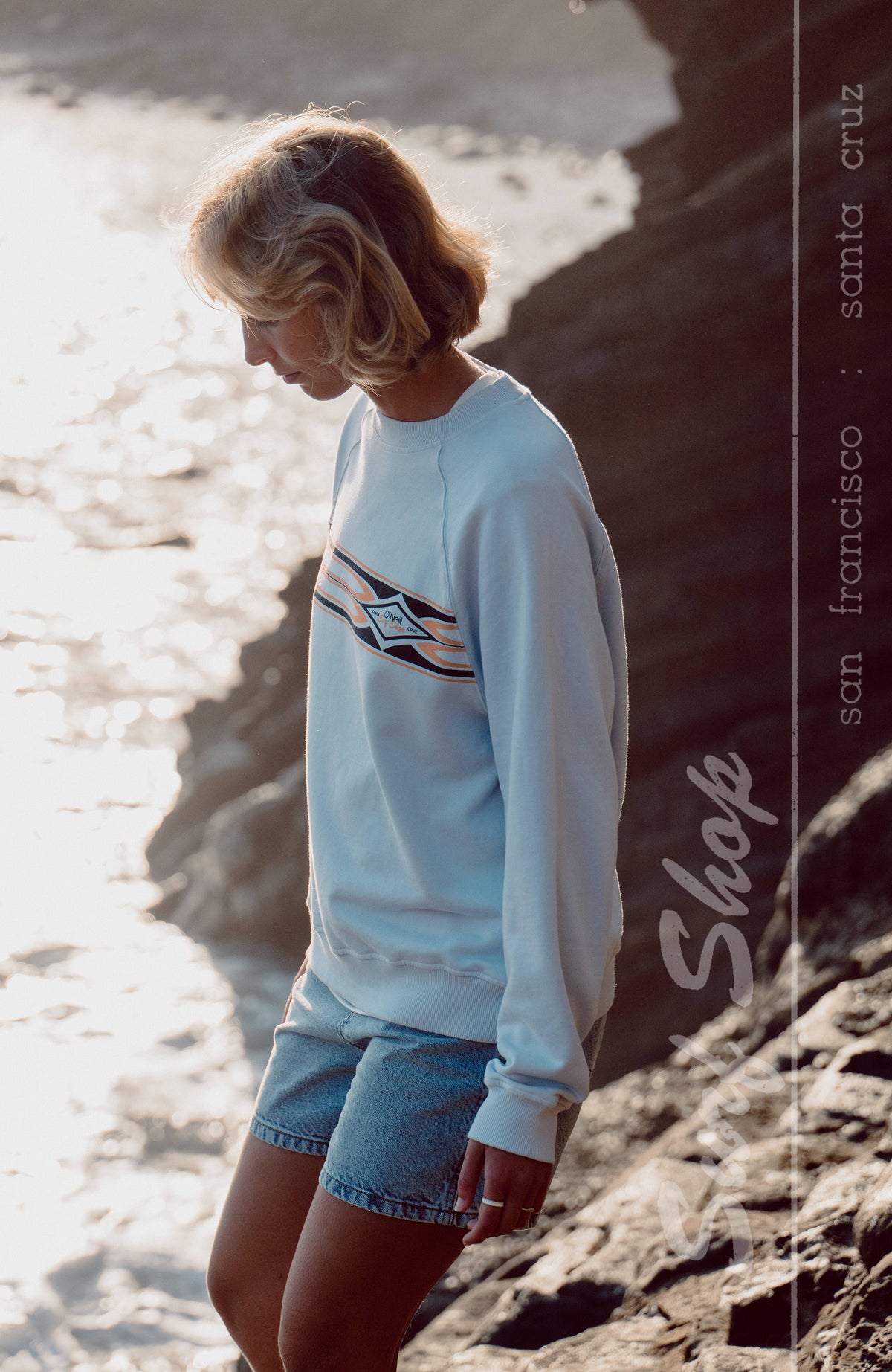 O'Riginals Crewneck Sweatshirt für Damen, blau mit Logo-Print, getragen an Felsen am Wasser