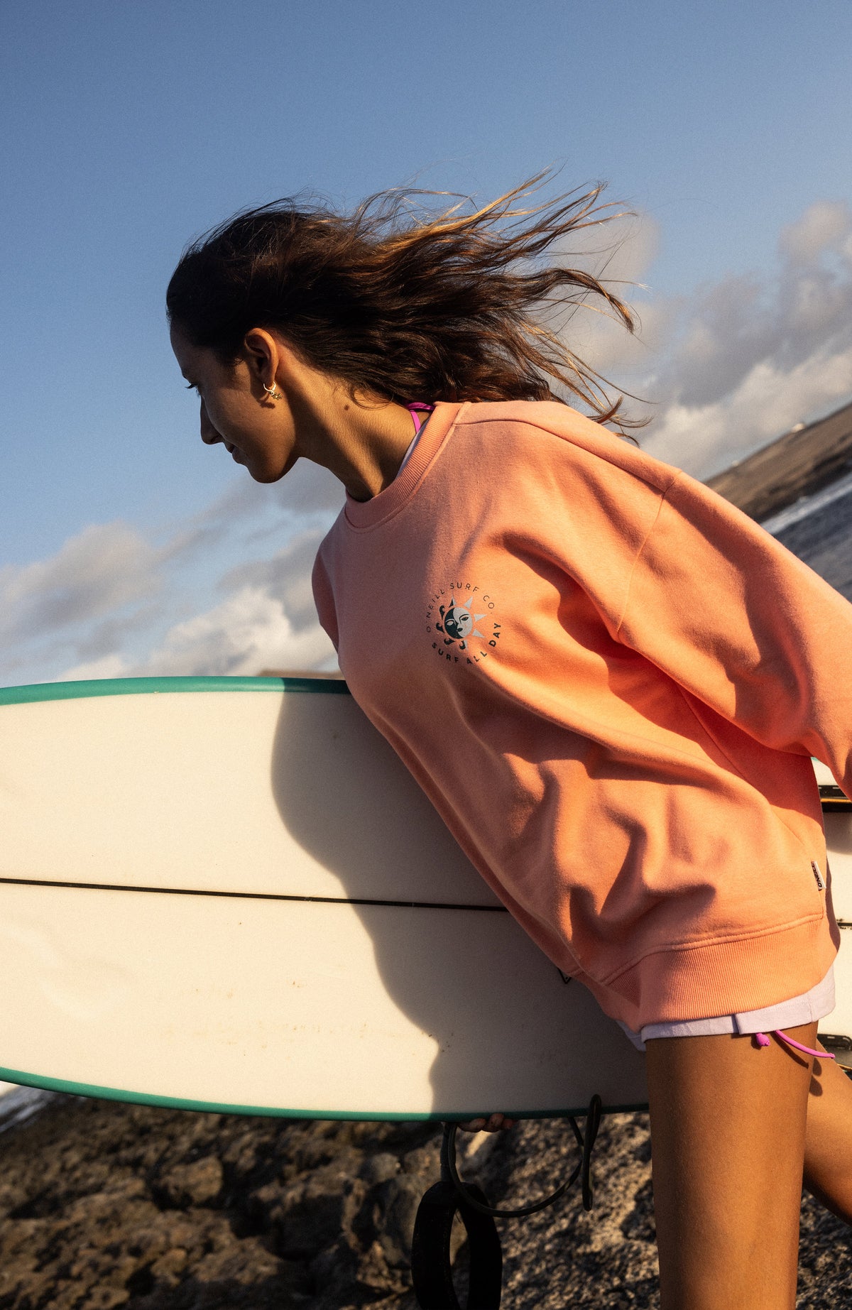 Summer Must-Have Crewneck Sweatshirt für Damen, orange, getragen am Model an der Küste mit Surfbrett