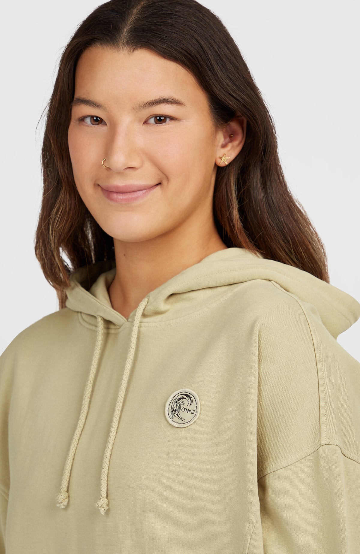 Nahaufnahme des beigen O'Riginals Badge Hoodie am Model mit runder Logo-Badge und Kordelzug