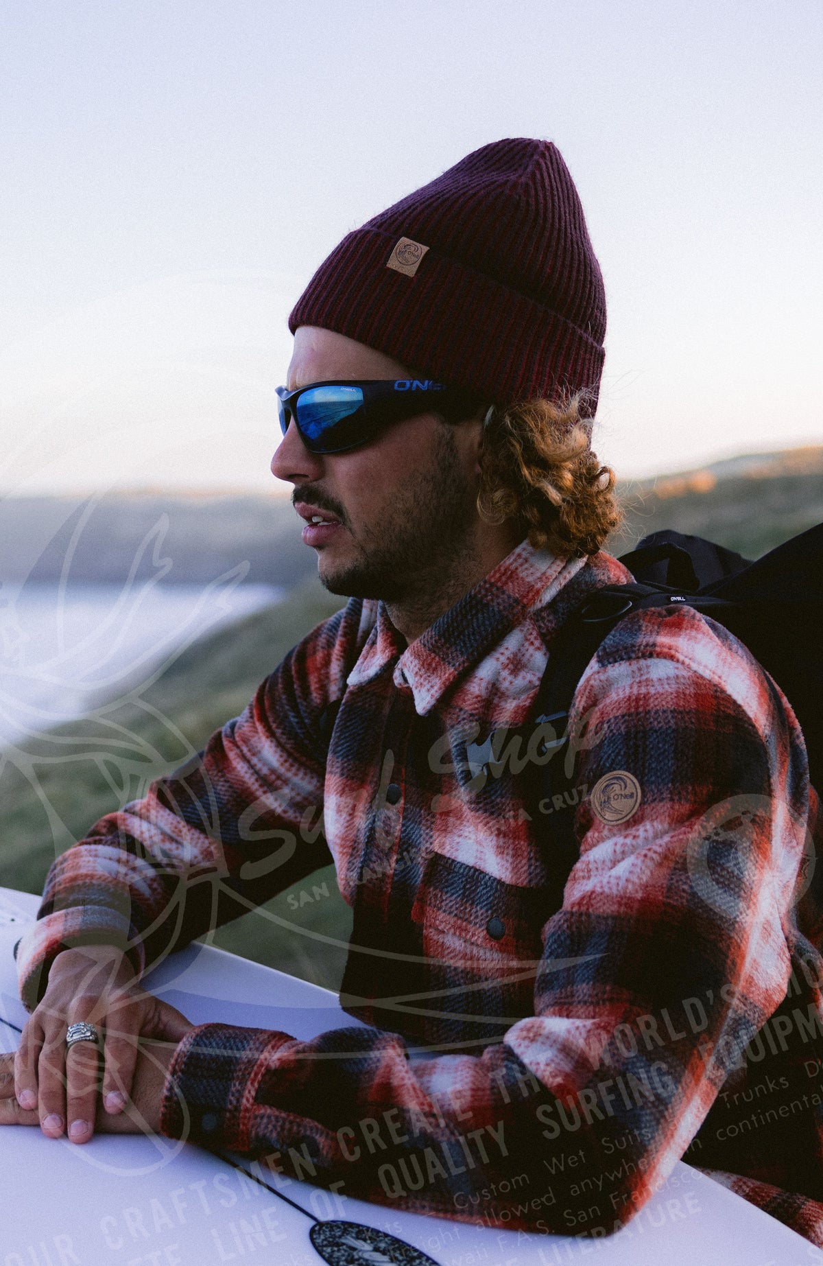 O'Riginals Beanie, orange, getragen am Model an der Küste mit Surfbrett