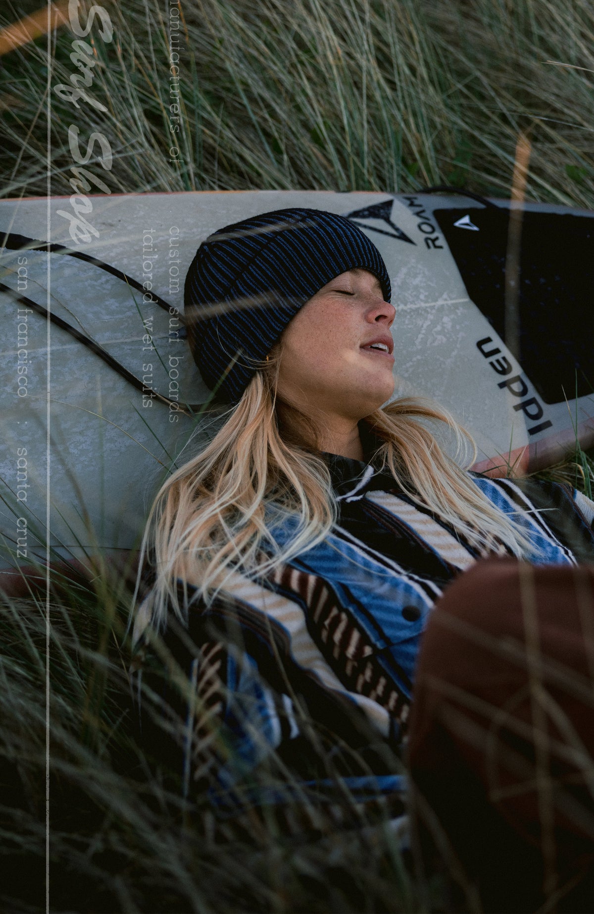 O'Riginals Beanie, blau, im Dünengras auf einem Surfbrett getragen am Model