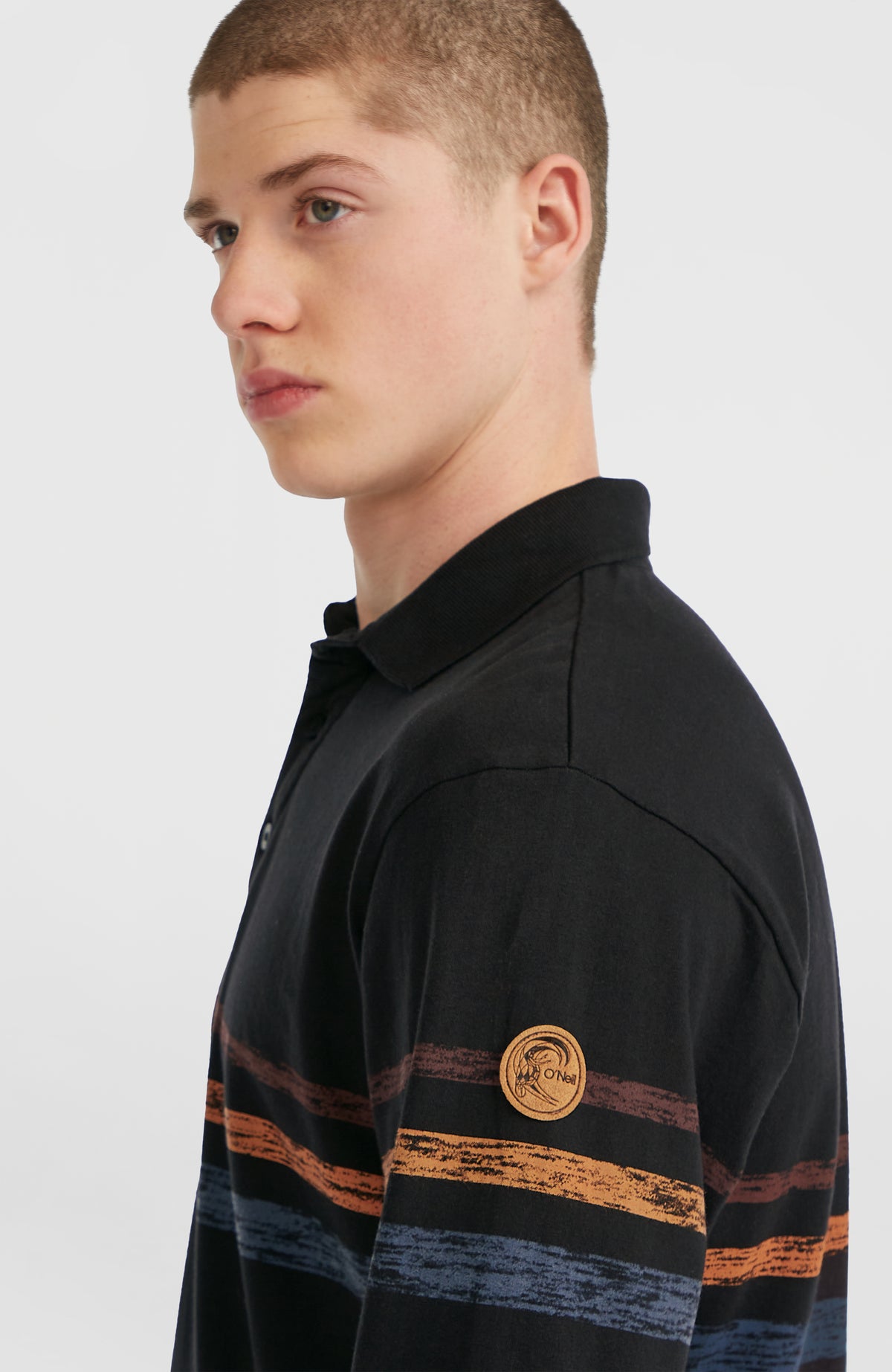 O'Riginals Poloshirt, schwarz mit Streifenmuster in Orange und Blau, Nahaufnahme am Model mit Logo-Patch am Ärmel