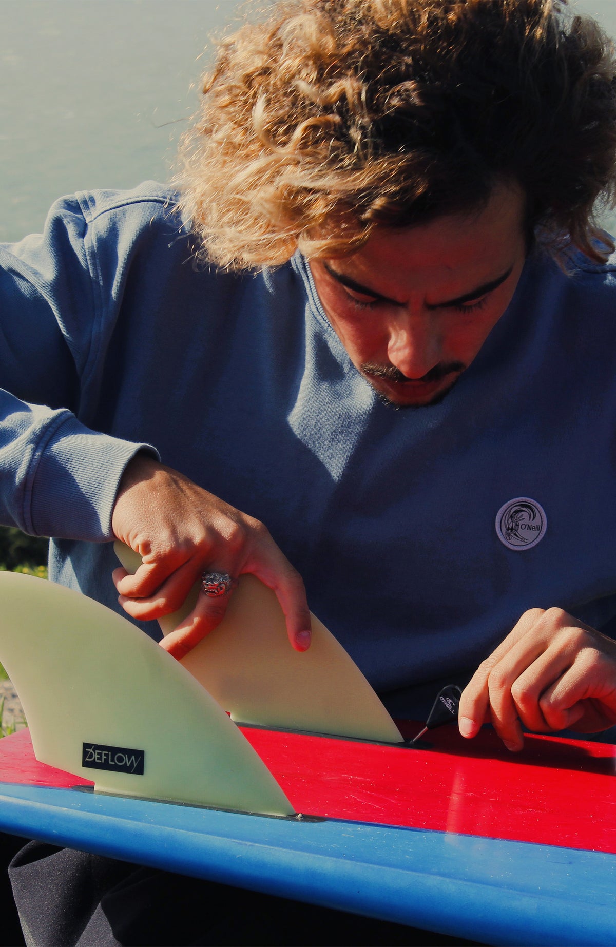 O'Riginals Crew Sweatshirt, blau, getragen am Model mit Surfbrett im Freien
