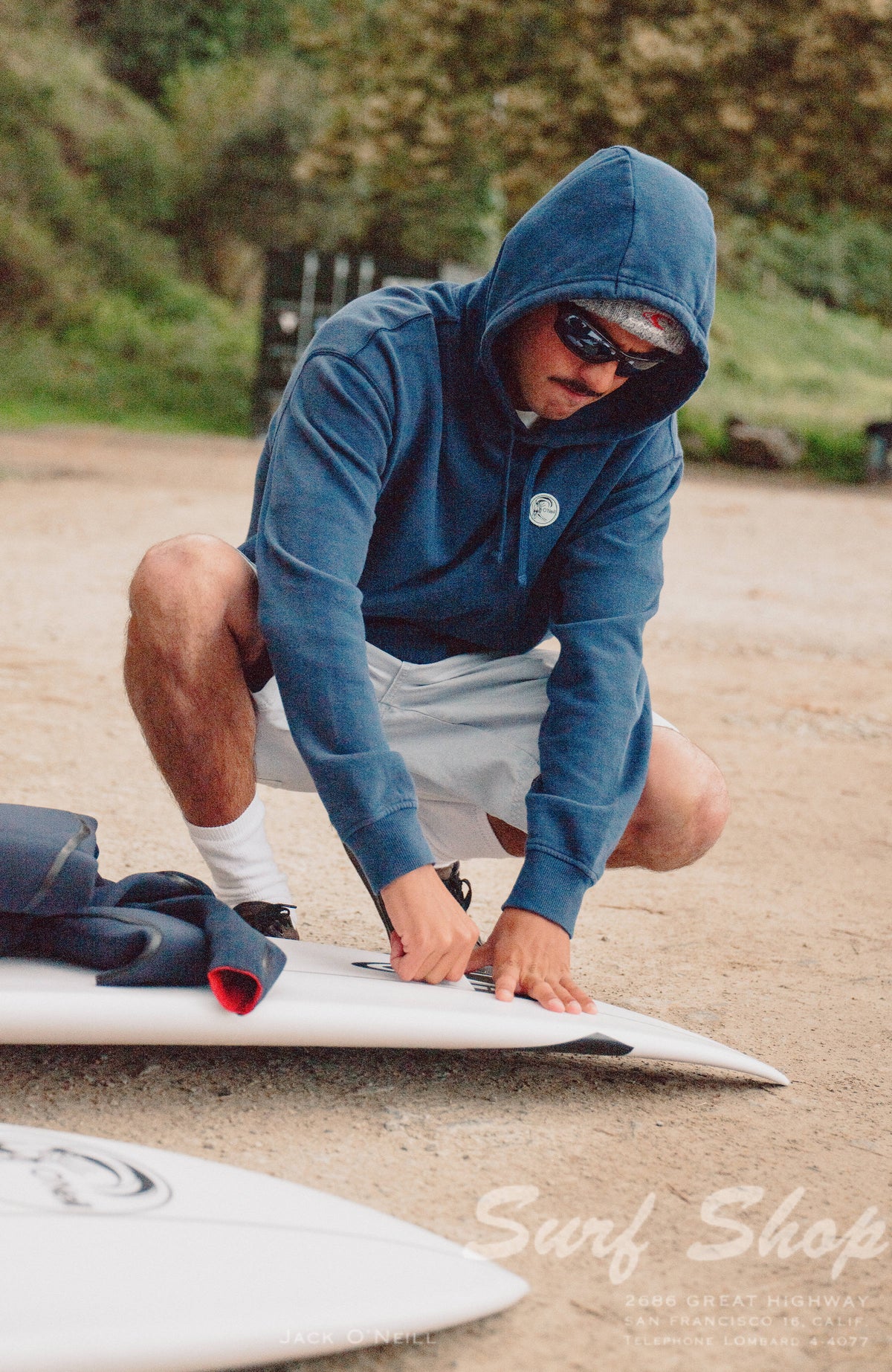 O'Riginals Hoodie für Herren, blau, am Strand mit Surfbrett, aufgesetzter Logo-Badge auf der Brust