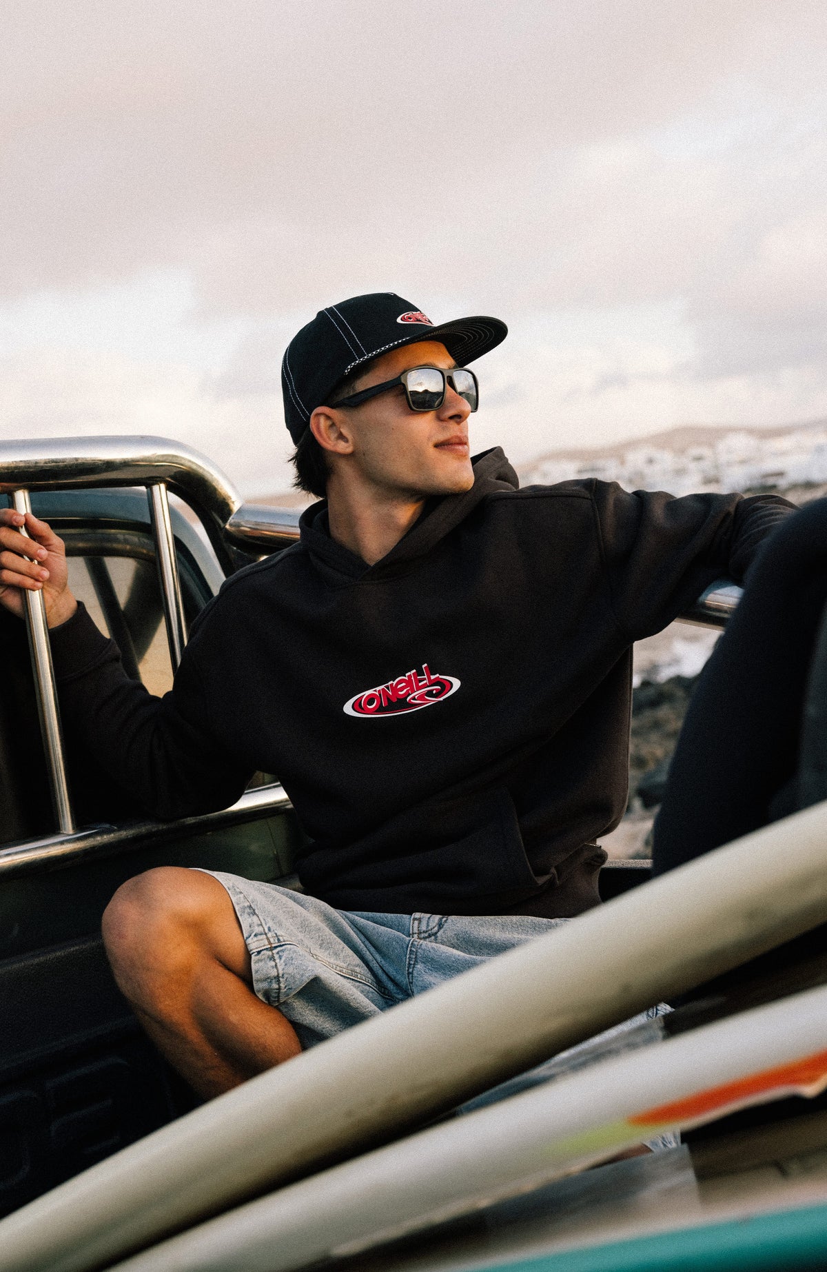 Essentials 90s Logo Hoodie, schwarz mit Logo-Print in Rot und Weiß, getragen am Model auf einer Ladefläche