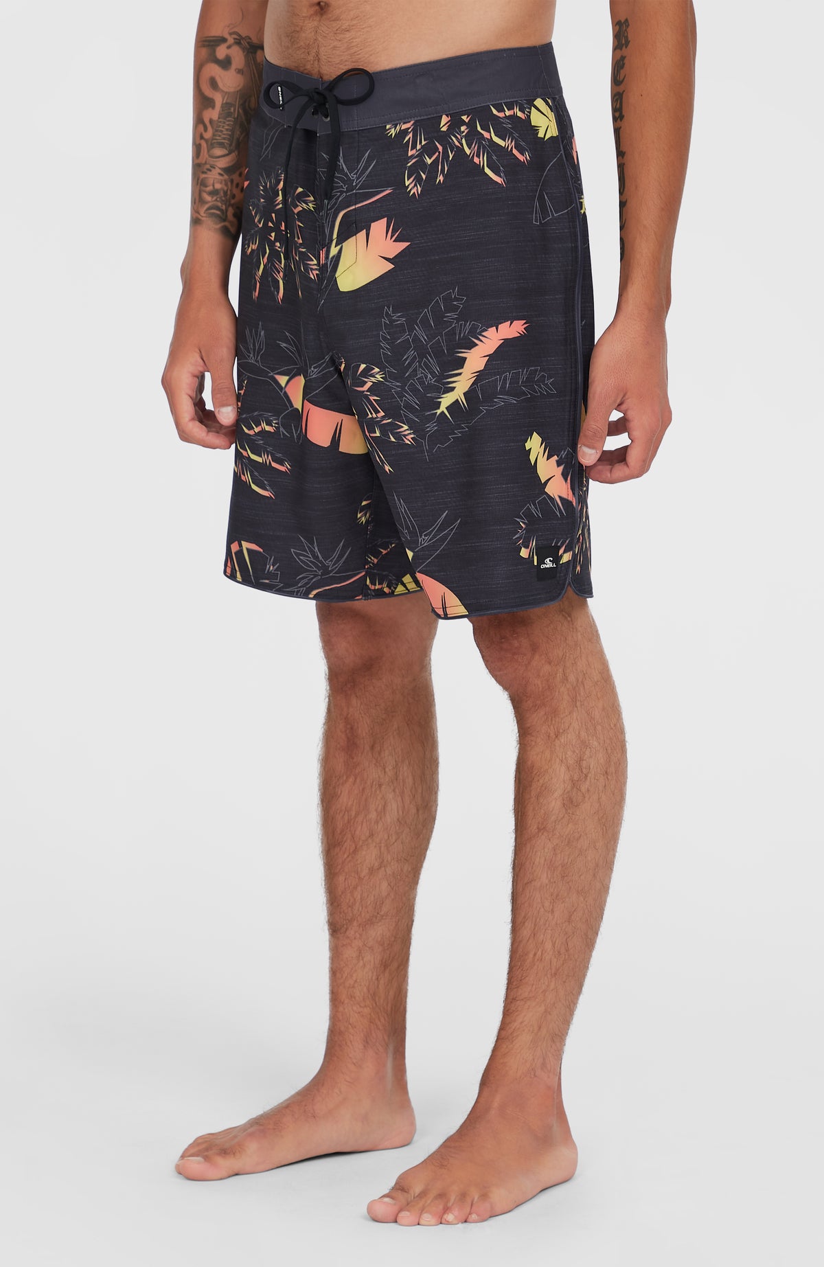 Hyperfreak Mysto Boardshorts, schwarz mit Palmenmuster in Orange und Gelb, schräge Seitenansicht mit Logo-Patch am Saum