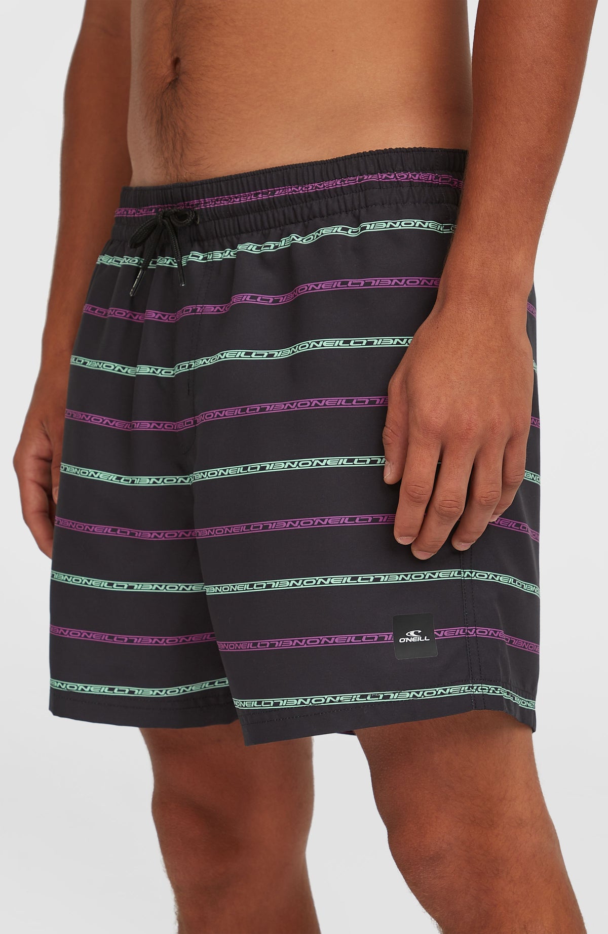 Nahaufnahme der schwarzen Cali First 15'' Badehose mit Streifenmuster in Lila und Türkis, Logo-Patch am Saum