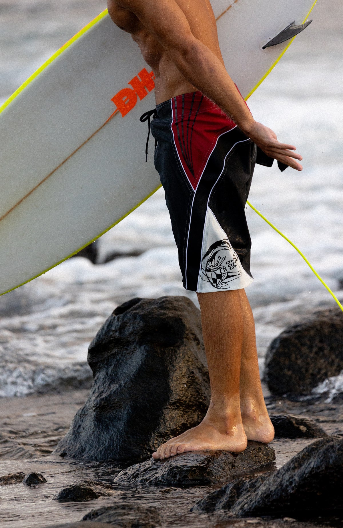 Superfreak Re-Issue Boardshorts, schwarz, Seitenansicht am Model am felsigen Meerufer mit Surfbrett
